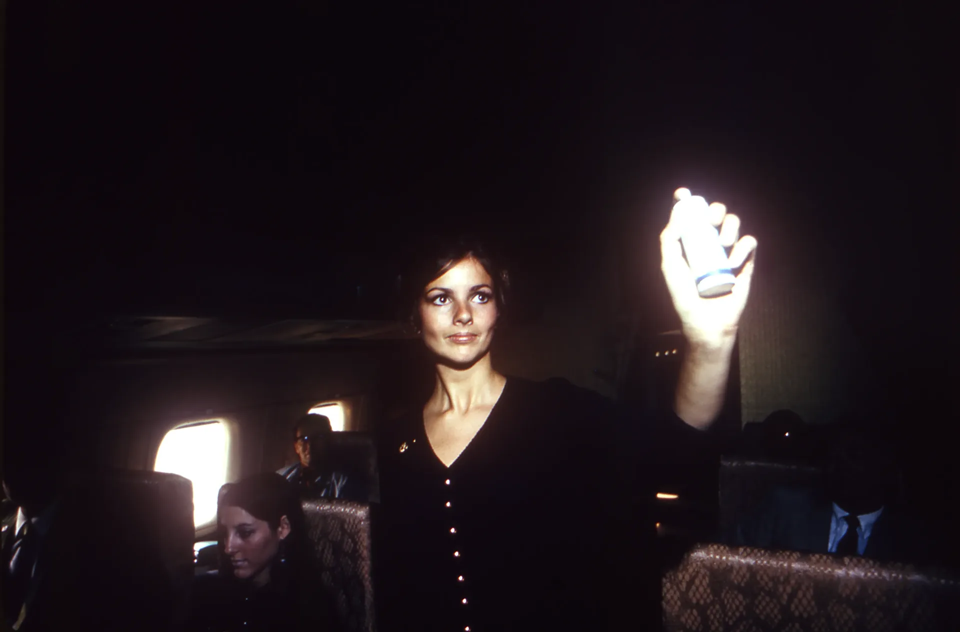 Flight attendant holding up an aerosol insecticide can in a dimly lit aircraft cabin with passengers seated, 1971.