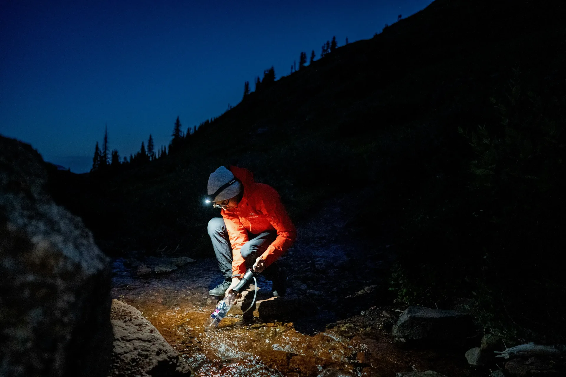 Hiker in a red jacket and headlamp crouching beside a mountain stream at dusk, using a portable water filter to fill a bottle