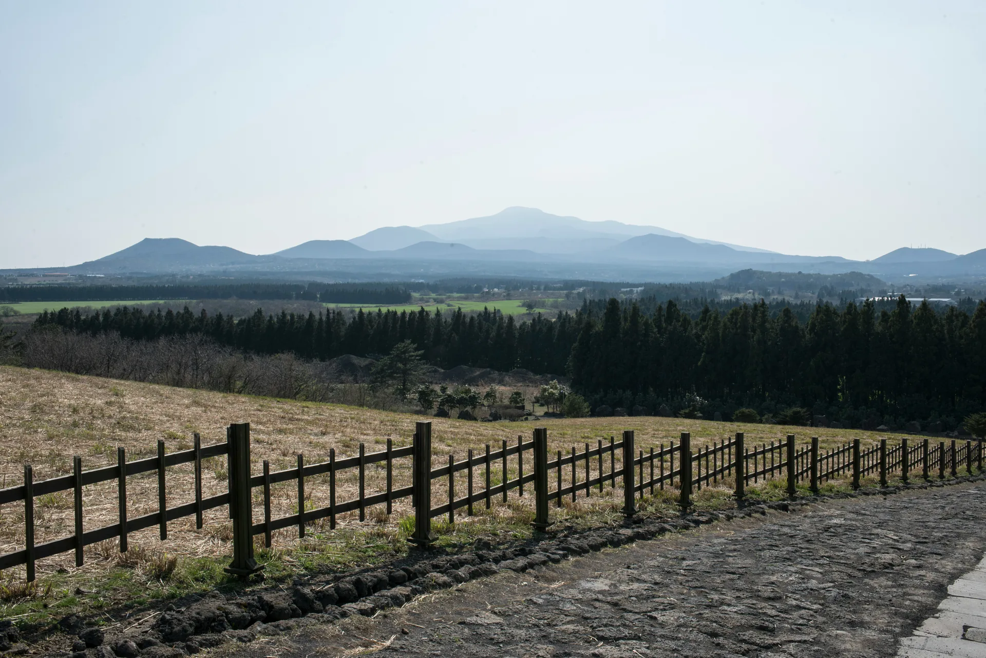 Wooden fence along dry farmland on Jeju Island, South Korea, with pine forest and hazy volcanic peaks in the distance