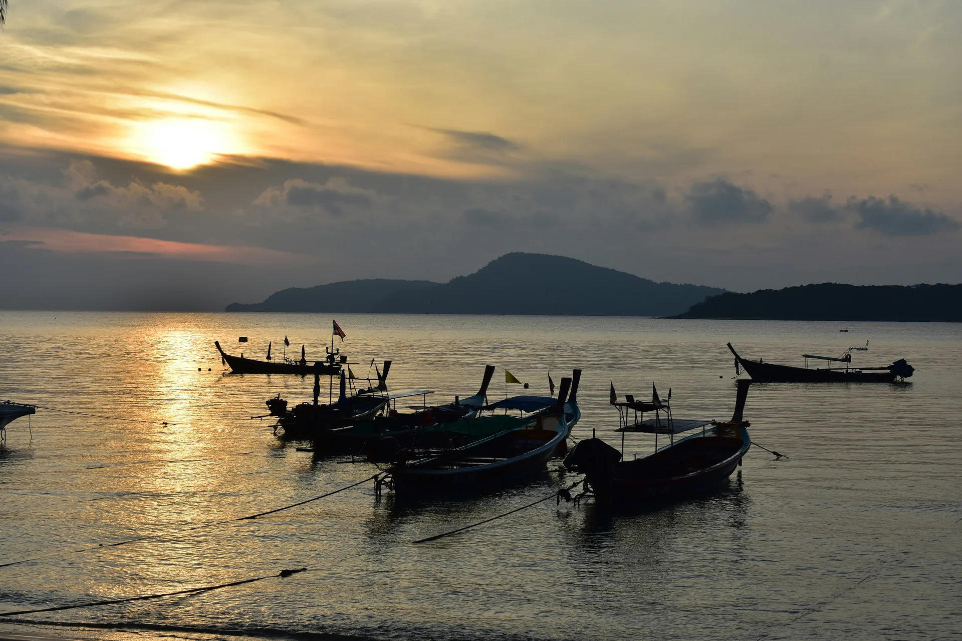 Longtail boats moored off Rawai Beach in Phuket, Thailand, at sunset with hills across the bay